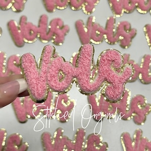 May include: Pink fluffy fabric patches with gold glitter trim spelling out the word "YOLO".