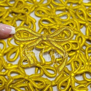 May include: A close-up of a pile of gold-colored fabric bows. The bows are all the same size and shape, and they are arranged in a random pattern. The bows are made of a soft, shiny fabric.