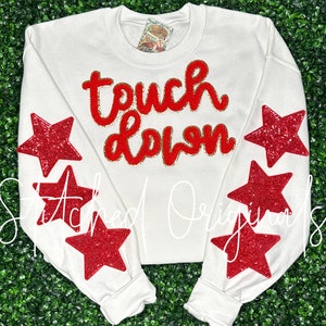 May include: White sweatshirt with the words "touch down" in red and gold glitter lettering. The sweatshirt has red glitter star appliques on the sleeves.