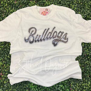 May include: A cream-colored short-sleeve t-shirt featuring the word Bulldogs in a cursive, rhinestone-encrusted font across the chest. A small rectangular tag with a graphic illustration is visible at the inner neckline of the garment.