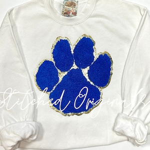 May include: White sweatshirt with a blue and gold paw print design. The paw print is made of fuzzy material and has a gold glitter outline.