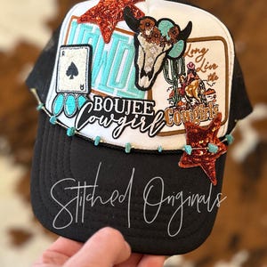 May include: Black and white trucker hat with a variety of colorful patches, including a turquoise skull with a floral crown, a turquoise and orange star, a turquoise and white patch that says "Long Live the Cowgirls", and a turquoise and white patch that says "Boujee Cowgirl".