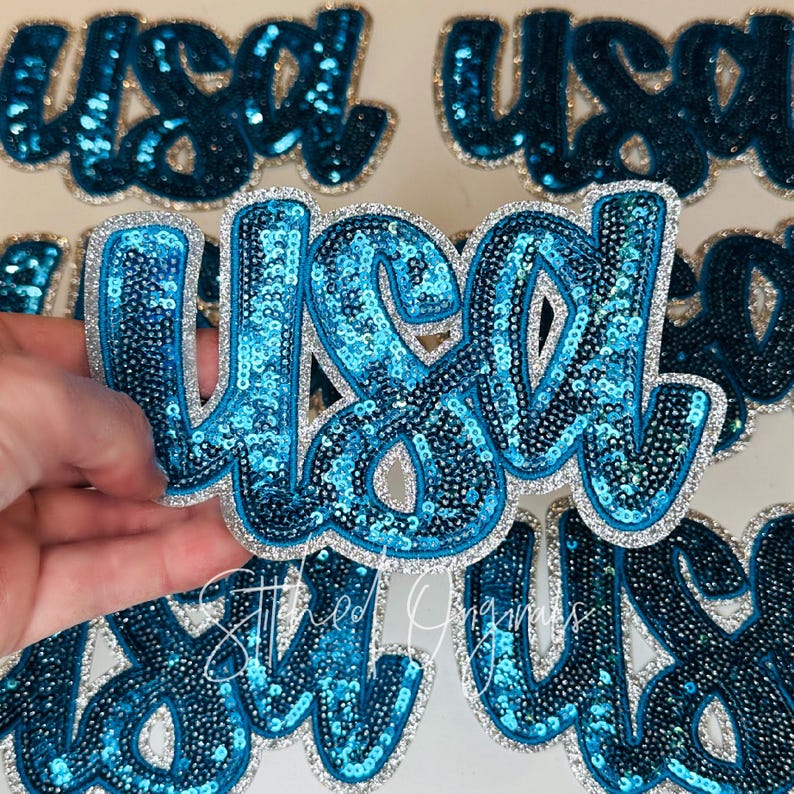 May include: A close-up of several sequined patches spelling "USA" in a cursive font. The patches are primarily turquoise blue with a silver glitter border. The patches are on a white surface.