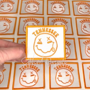 May include: White square patch with orange embroidered border and design. The patch features the word "Tennessee" above a smiley face with crossed-out eyes and a protruding tongue. The background shows multiple similar patches.