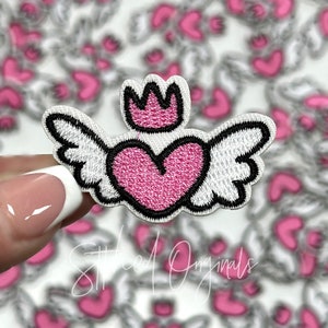 May include: Pink heart with white wings and a pink crown on top. The heart is outlined in black. The patch is iron-on and perfect for adding a touch of whimsy to any project.