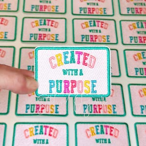 May include: A rectangular patch with the words "CREATED WITH A PURPOSE" embroidered in colorful thread. The patch has a white background with a teal border. The text is in a rainbow of colors.