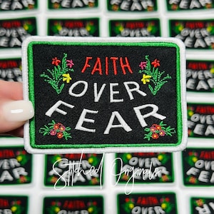 May include: A black fabric patch with white text that reads "FAITH OVER FEAR" in a bold font. The patch is outlined in green and white and has small embroidered flowers in red, yellow, and green.