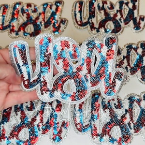 May include: A collection of sequined patches spelling out "USA" in a cursive font. The patches feature red, white, and blue sequins, with a silver glitter border. The patches are designed for decorative purposes.