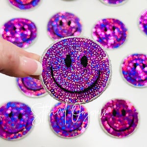 May include: Purple and pink sequined smiley face iron-on patches. The patches are about 2 inches in diameter and have a white outline.