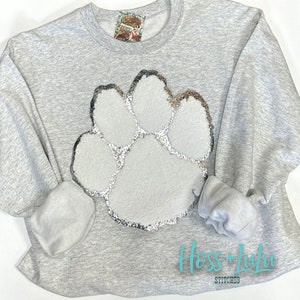 May include: Gray sweatshirt with a large white glitter paw print design. The sweatshirt has the text "Hoss + Lulu Stitched" on the bottom.