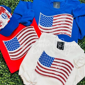 May include: Three sweatshirts with a sequined American flag design. The sweatshirts are in red, white, and blue.