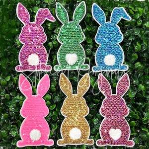 May include: Six sparkly sequin bunny rabbit patches in various colors, including pink, green, blue, gold, and purple. Each patch has a white felt tail and a white heart in the center.