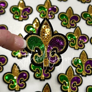 May include: A collection of fleur-de-lis patches in gold, green, and purple sequins, with black borders. The patches are in the shape of a stylized lily, a symbol of royalty. The text "Stitched Originals" is visible on the image.
