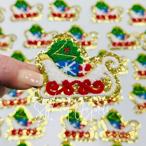 May include: Gold glitter iron-on patch of a white sleigh with a green Christmas tree, red and white candy canes, and a blue snowflake. The sleigh is outlined in gold and has a red base.