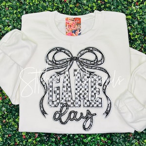 May include: White sweatshirt with a black and white checkered "GAME DAY" design. The design includes a bow and is outlined with silver sequins. The sweatshirt has long sleeves and a crew neck.