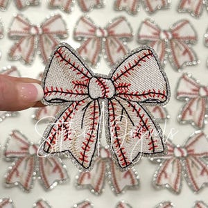 May include: A white and red baseball-themed bow with a sparkly silver trim. The bow is made of fabric and has a stitched baseball design.