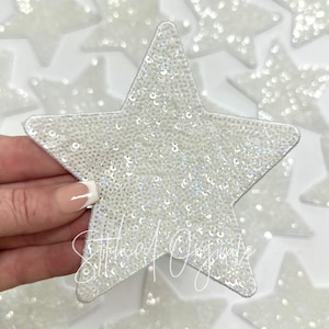 May include: A white star-shaped patch covered in iridescent sequins. The patch is approximately 4 inches in diameter.