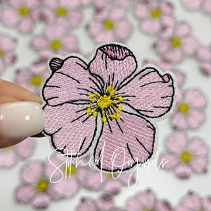 May include: Embroidered pink flower patch with black outline and yellow center. The patch has a white border and is held in front of a blurred background of similar patches. The text "Stitched Originals" is written across the bottom of the image.