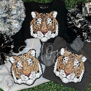 May include: Black, gray, and light gray sweatshirts with a sequined tiger face design. The tiger face features gold, brown, and white sequins with a red nose. The sweatshirts are displayed with silver and black pom-poms.