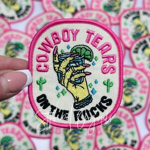May include: Pink and white embroidered patch with the text "Cowboy Tears on the Rocks" and a cartoon hand holding a shot glass with a lime wedge.