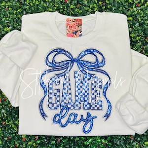 May include: Cream-colored sweatshirt with a blue bow and the words "GAME DAY" in a blue checkered pattern. The sweatshirt has long sleeves and a crew neck. The design is embellished with glitter.