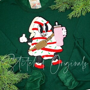 May include: Green sweatshirt with a cartoon Christmas tree wearing a red and white striped outfit and holding a pink mug. The tree has googly eyes and a toothy grin. The text "Stitched Originals" is visible at the bottom of the sweatshirt.