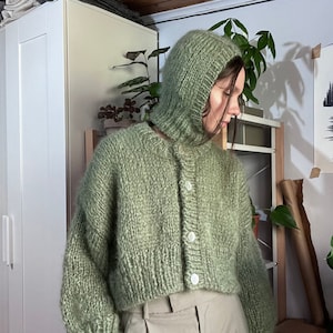 May include: A sage green knitted cardigan with a hood and button closure. The cardigan is cropped and has a relaxed fit.