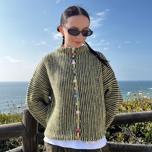 Peut inclure: Un cardigan rayé jaune et noir avec un col montant et des boutons colorés. Le cardigan est boutonné et porté sur une chemise blanche. La personne porte des lunettes de soleil et se tient dehors avec vue sur l'océan.