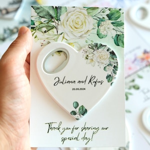 Può includere: Un apribottiglie bianco a forma di cuore con un design floreale e i nomi "Juliana e Rufus" e la data "20.05.2024". La carta recita: "Grazie per aver condiviso il nostro giorno speciale!"