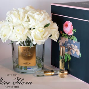May include: A bouquet of white roses in a clear glass vase with a gold label that says "Miss Flora Flower Shop". The vase is sitting on a table next to a black gift box with a pink rose design and a small bottle of perfume.
