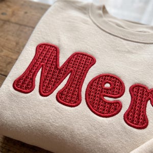 Puede incluir: Una sudadera de cuello redondo color crema con la palabra "Mem" bordada en letras rojas grandes y llamativas. Las letras tienen una textura de punto trenzado en relieve que destaca sobre el tejido liso de la prenda.