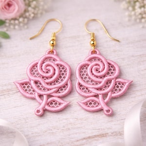 May include: A pair of pink rose-shaped earrings. The earrings feature a delicate, open-weave design, with gold-coloured ear wires. The rose design includes petals and leaves, creating a floral theme. The earrings are displayed on a light-coloured surface.