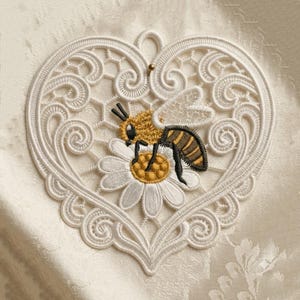 Puede incluir: Aplique de encaje blanco en forma de coraz&oacute;n con un dise&ntilde;o detallado. Una abeja bordada en amarillo, negro y marr&oacute;n est&aacute; posada sobre una margarita blanca con un centro amarillo. El aplique tiene un peque&ntilde;o lazo en la parte superior para colgar.