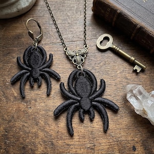 Op de afbeelding: Een sieradenset bestaande uit een ketting en een enkele oorbel, beide voorzien van zwarte geborduurde spinnenmotieven. De ketting bevat een bronskleurige ketting met een klein bijvormig verbindingsstuk aan de spinnenhanger.