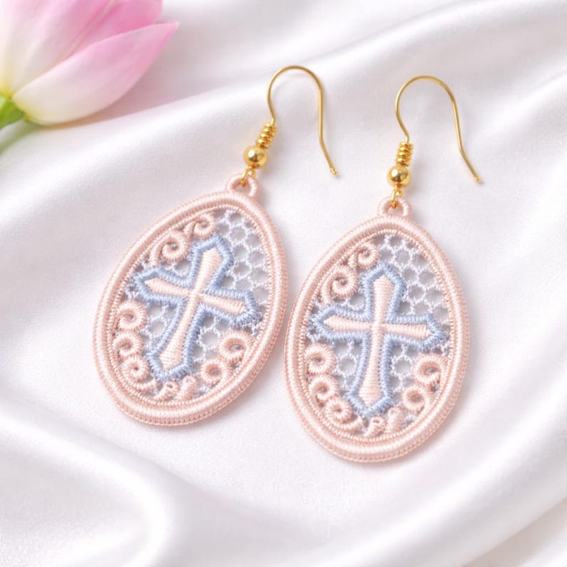 May include: A pair of oval-shaped earrings with a cross design. The earrings feature a light pink border with a light blue cross and white filigree. The hooks are gold-colored.