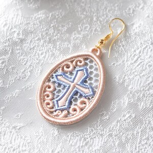 May include: A single gold-tone earring with an oval, openwork design. The earring features a cross design in light blue and pink embroidery, framed by a pink border and gold-tone hook. The background is a white, textured fabric.