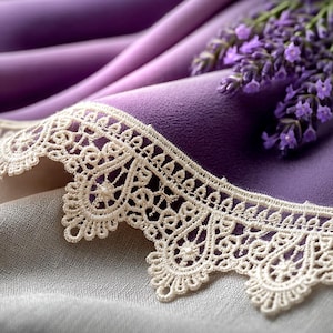 Puede incluir: Primer plano de una tela morada con un delicado ribete de encaje color crema. Una ramita de flores de lavanda descansa sobre la tela, añadiendo un toque de belleza natural. Los suaves pliegues de la tela crean una sensación de profundidad y textura.