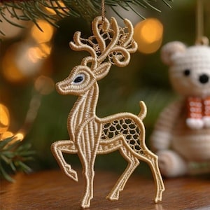 May include: A gold-colored, embroidered reindeer Christmas ornament. The reindeer has detailed antlers and a delicate openwork design on its body. It is hanging from a gold string, with a blurred background of Christmas tree branches and lights.