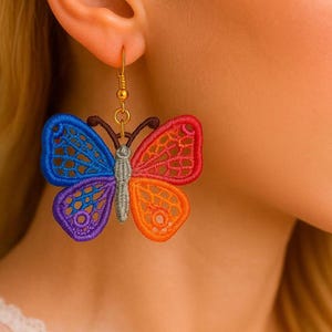 Pode incluir: Um brinco de borboleta colorido com um gancho dourado. O design da borboleta apresenta secções azuis, roxas, laranja e vermelhas. O brinco é feito de um material delicado e vazado.