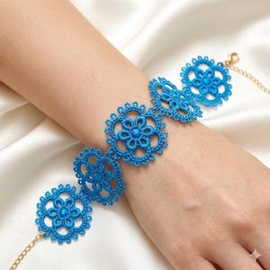 Puede incluir: Una pulsera de encaje azul brillante que presenta una serie de motivos florales circulares interconectados. El intrincado trabajo de hilo está unido por una fina cadena de metal dorado con un cierre de mosquetón, creando un accesorio.