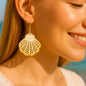 May include: Gold-toned seashell earrings with a cream-colored top and a detailed, openwork design. The earrings hang from a delicate gold hook. The earrings are a beach-themed accessory.