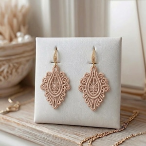 May include: A pair of light pink, ornate lace earrings with gold-coloured hooks. The earrings are displayed on a white stand, showcasing their intricate, symmetrical design. The earrings have a teardrop shape with detailed patterns.