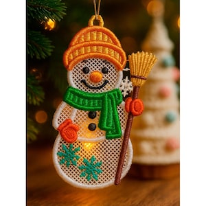Könnte beinhalten: Gestickter Schneemann-Weihnachtsschmuck mit orangefarbener Mütze, grünem Schal und orangefarbenen Fäustlingen. Der Schneemann hält einen Besen und hat ein lächelndes Gesicht mit schwarzen Knopfaugen. Zwei grüne Schneeflocken.