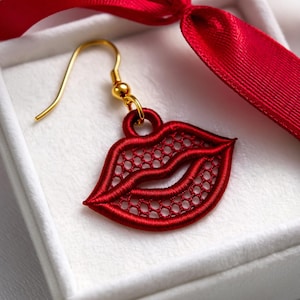 Puede incluir: Un solo pendiente rojo bordado con forma de labios con un gancho dorado. El pendiente se muestra en una caja blanca con una cinta roja. Los labios están intrincadamente detallados con un patrón similar a una malla.