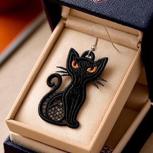 May include: A black cat-shaped earring with orange eyes and delicate lace detailing, displayed in a navy blue jewellery box. The earring has a silver hook and a detailed design, ideal for cat enthusiasts.