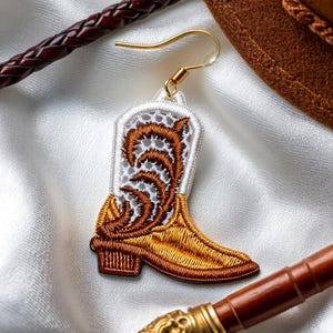 May include: Embroidered cowboy boot earring with gold-colored hook. The boot features a white and brown design with gold and brown embroidery. The earring is set against a white satin background, with a brown leather strap and a brown hat in the background.
