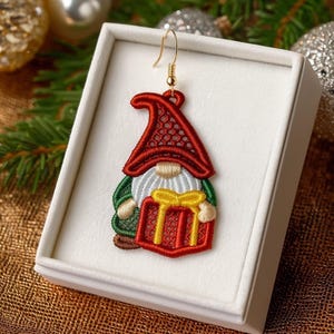 Op de afbeelding: Geborduurde kabouteroorbel met een rode hoed, witte baard en groene jas, met een rood cadeau met een gele strik. De oorbel heeft een goudkleurige haak en wordt gepresenteerd in een witte doos.