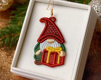 May include: Embroidered gnome earring with a red hat, white beard, and green jacket, holding a red gift with a yellow bow. The earring has a gold-colored hook and is displayed in a white box.
