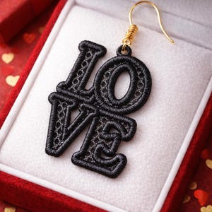May include: A black embroidered earring spelling out "LOVE" with a gold-colored hook, presented in a red jewelry box with a white interior. The background features a red surface with heart-shaped confetti.
