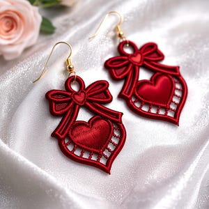 May include: A pair of red heart-shaped earrings with gold-colored hooks. Each earring features a bow at the top and a heart in the center, with a delicate, openwork design. The earrings are displayed on a white, silky surface.